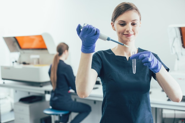 Positive laboratory technician using a manual pipette and smiling while dropping sample into a test tube in her hand. Colleague at the table on a background