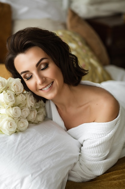 Positive lady in bathrobe with makeup on her eyes smiling while relaxing on the bed with a bouquet of white roses.