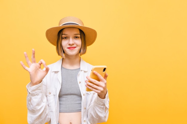 Positive lady in a hat with a phone in hand stands on a yellow background looks in camera