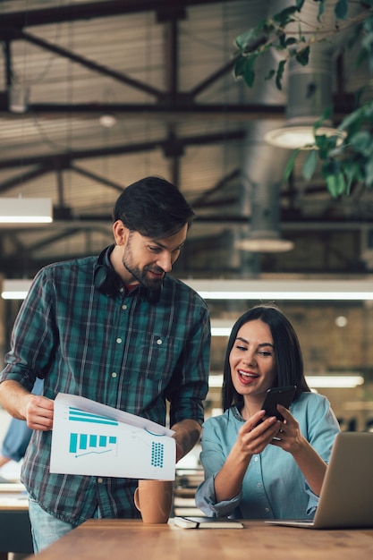 Positive lady sitting with a gadget and a friendly colleague standing by her side with graphics