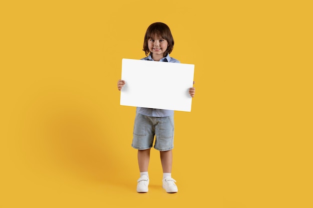 Positive little boy holding blank card and smiling to camera posing over orange studio background empty space for text