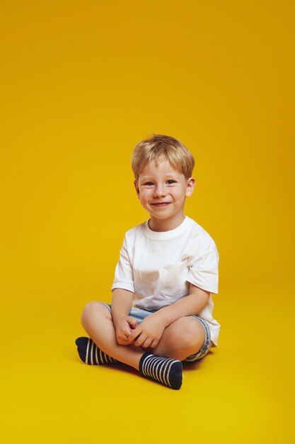 写真 白いトレンディなtシャツを着たポジティブな小さな男の子が笑顔で横足で座ってカメラを見ている