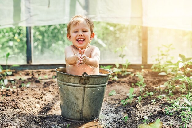 Positive little boy sits in old metal bucket of warm water on kitchen garden bed in old greenhouse