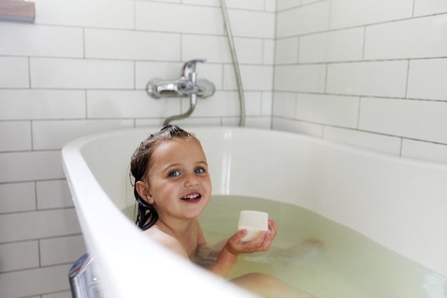 Positive little girl sitting in water of bath with bar soap while washing in bathtub 