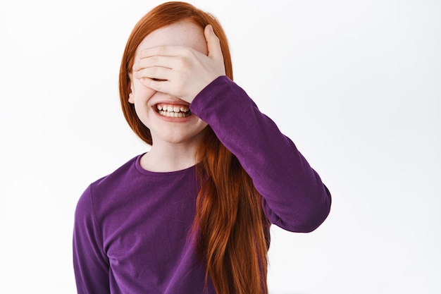 Positive little girl with red hair and freckles smiling, covering eyes and waiting for surprise, standing excited against white background