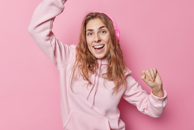 Positive long haired woman with glad expression dances carefree moves with rhythm of music wears stereo headphones on ears casual sweatshirt isolated over pink background People and happiness