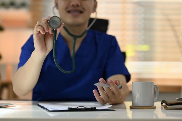 Positive male doctor in uniform holding stethoscope and smiling at camera