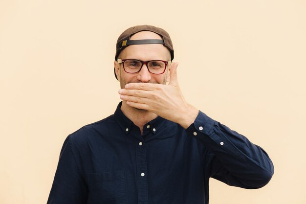 positive male model covers mouth as giggles at funny joke wears spectacles cap and elegant shirt expresses good emotions poses against beige background people emotions lifestyle concept
