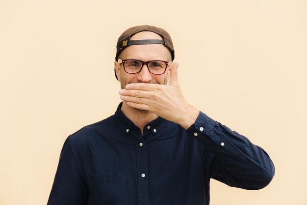 Positive male model covers mouth as giggles at funny joke wears spectacles cap and elegant shirt expresses good emotions poses against beige background People emotions lifestyle concept