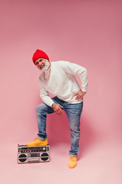 Positive man in jeans and sweater posing with record player on pink background grey haired guy in orange hat and yellow sneakers smiling