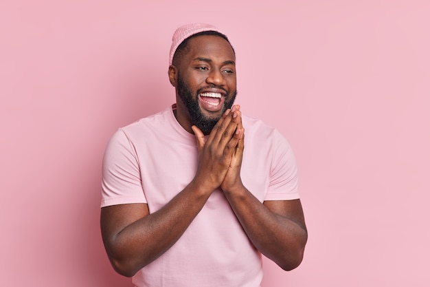 긍정적 인 남자가 손바닥을 함께 유지하는 것은 좋은 일에 대한 믿음을 가지고 있습니다 즐겁고 행복한 미소가 캐주얼 한 옷을 입고 광범위하게 옷을 입고 분홍색 벽에 고립 된 좋은 일이 있다고 믿습니다.