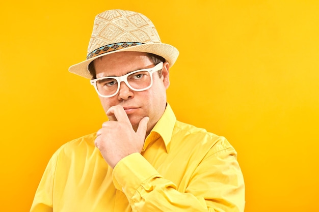 Positive man in panama hat holding chin thinks about vacation doubts makes decision isolated on yellow studio background