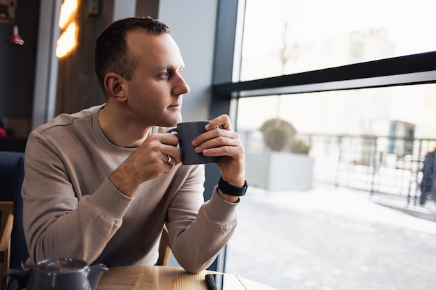 A positive man sits in a cafe at a table, he drinks aromatic coffee. Smiling guy in casual clothes sits in a cafe and drinks morning coffee