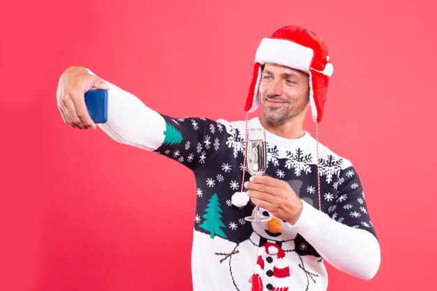 赤い背景、クリスマスにシャンパンを飲みながらスマートフォンでselfieを作る冬のセーターと帽子のポジティブな男。