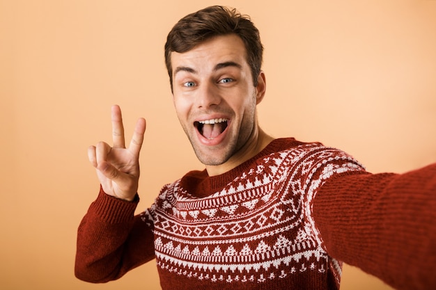 positive man with bristle wearing knitted sweater laughing and showing peace sign while taking selfie photo, isolated over beige wall