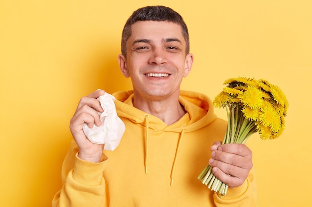 アレルギー症状があるが春を楽しんでいるタンポポの花束を保持している黄色の背景の上に孤立したポーズをとってカジュアルなスタイルのパーカーを着てハンカチを持つポジティブな男