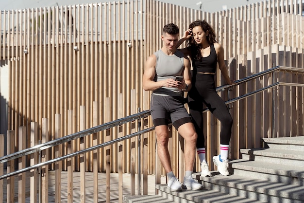 Positive man and woman resting after hard street workout session embracing using smartphone