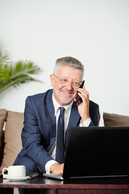 Positive mature businessman calling on phone and checking documents on laptop screen