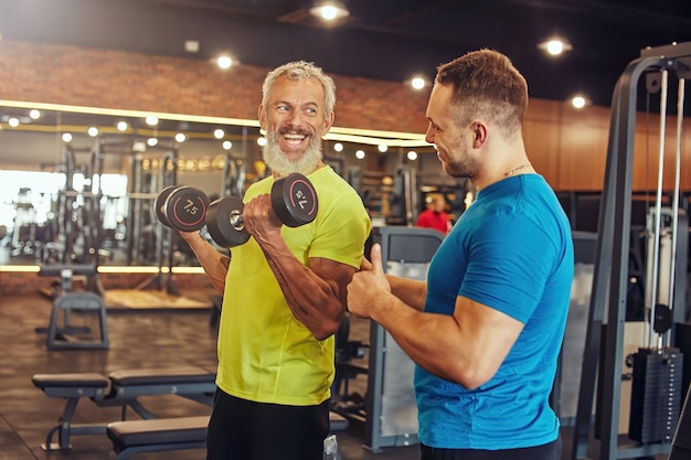 Uomo maturo positivo in abiti sportivi che fa esercizi con i pesi con il suo giovane personal trainer che lavora