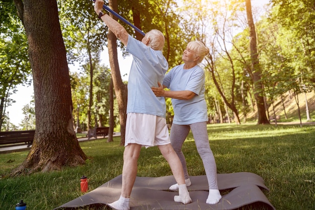 Positive mature woman supports back of senior man doing exercises with bar in park