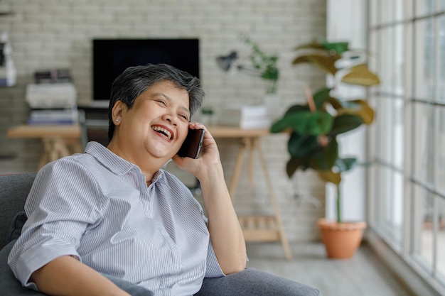 긍정적인 중년 아시아 여성은 소파에 앉아 현대적인 거실에서 스마트폰 대화를 하면서 웃고 멀리 바라보고 있습니다.