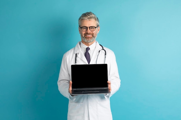 Positive middle aged doctor showing laptop with black screen