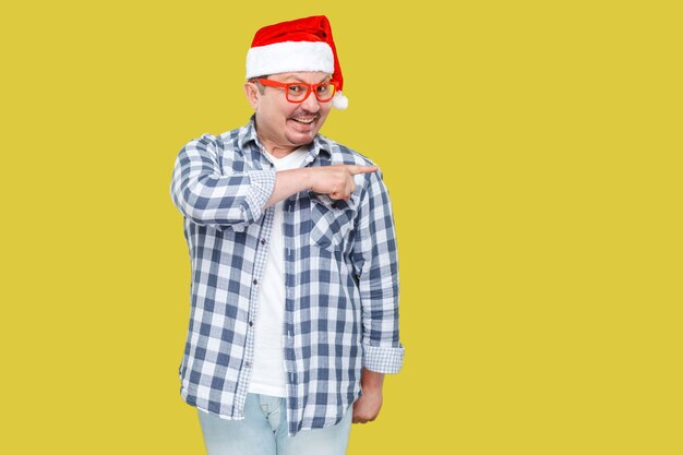 Positive middle aged man in red cap, eyeglasses and checkered shirt standing and pointing finger to empty copy space and toothy smiling, looking at camera. Studio shot, isolated on yellow background