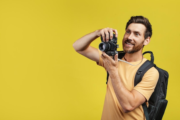 Positive middle aged man with backpack taking picture on professional DSLR camera over yellow background free space