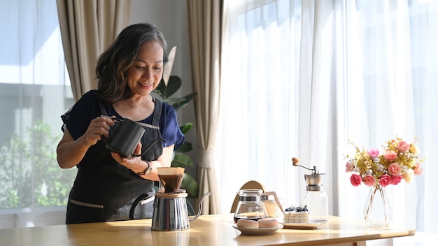 Positive middle aged standing at kitchen table brewing drip coffee and enjoys her morning