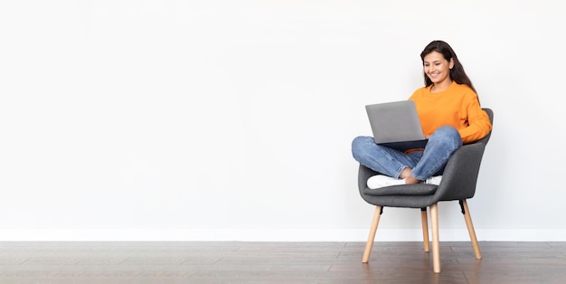 Positive middle eastern young lady using laptop sitting in armchair