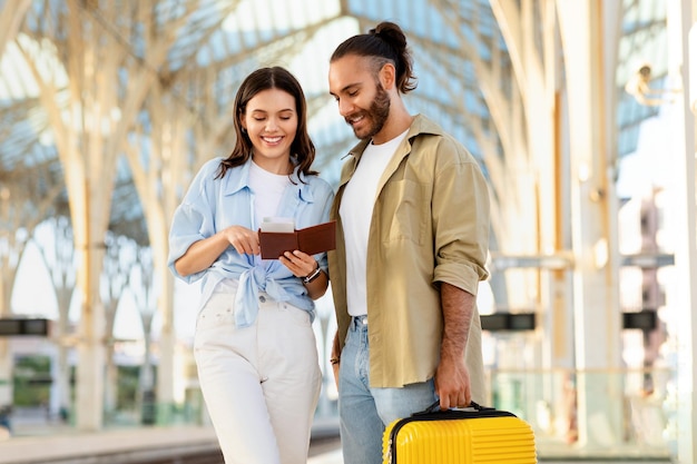 Positive millennial caucasian couple with suitcase looks at passport at train station