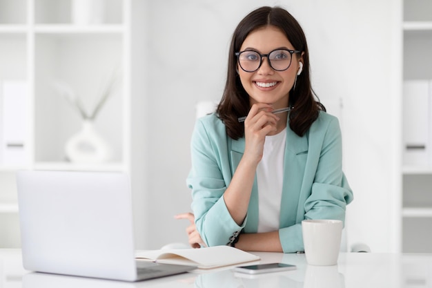 Positive millennial caucasian lady in suit and glasses at workplace with laptop enjoy coffee