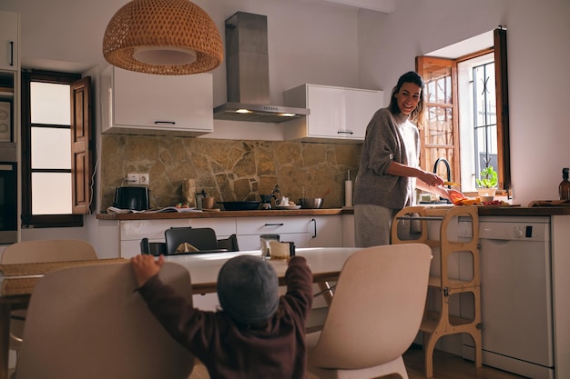 positive mom preparing food for small boy standing near table and chairs in kitchen