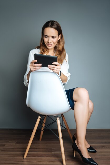 Positive mood. Attractive delighted nice woman holding a tablet and using it while resting on the chair