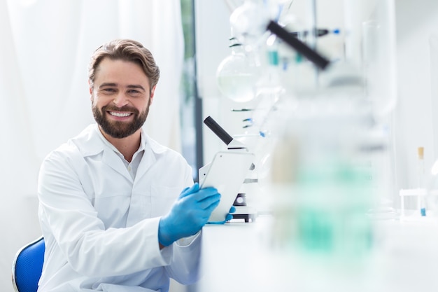 Positive mood. Cheerful nice male scientist sitting on the chair and smiling to you while being happy about his job in the lab