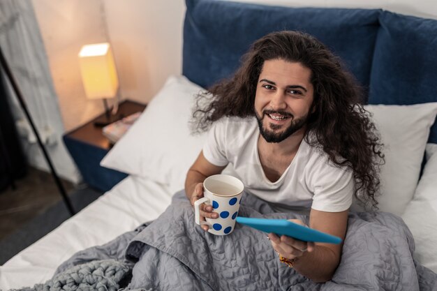 Positive mood. Cheerful nice man smiling to you while holding his tablet