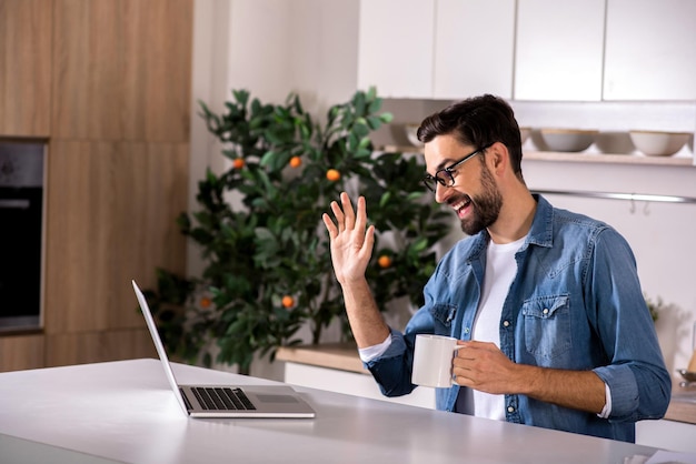 Positive mood Cheerful young man sitting in the kitchen and using his laptop