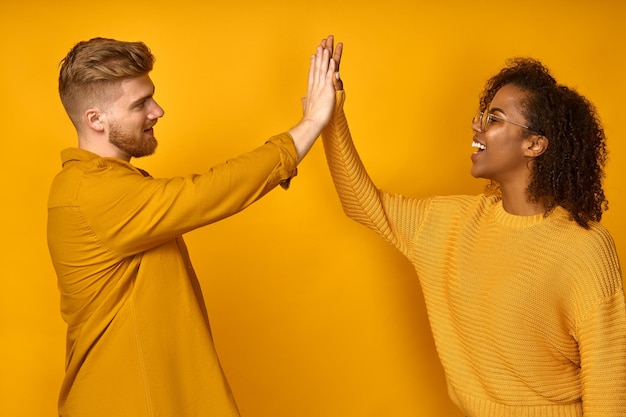 Positive multicultural couple give high five agree to work as team stand sideways on yellow wall