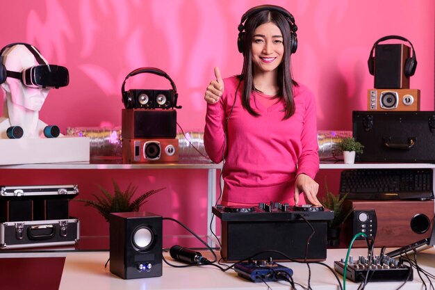 Positive musician standing at dj table doing thumbs up gesture while mixing electronic music and techno using mixer console. Asian artist playing stereo sounds with electronics and microphone