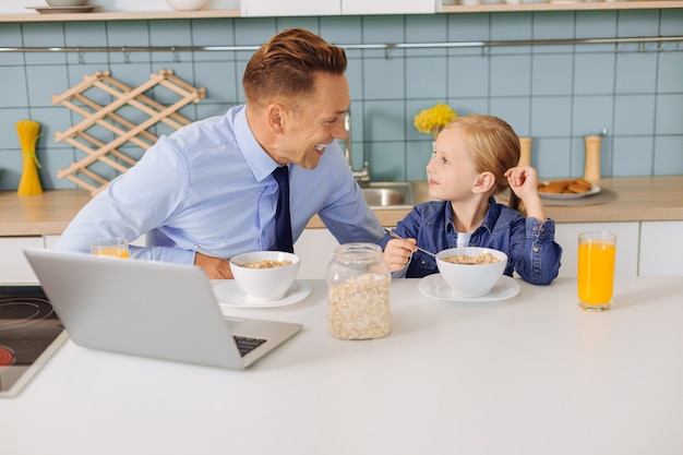 Positive nice blonde girl sitting at the kitchen table and looking at her father while eating food