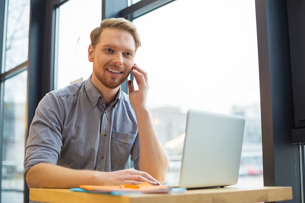 Positive nice handsome man holding his smartphone and making a call while sitting at the table in the cafe