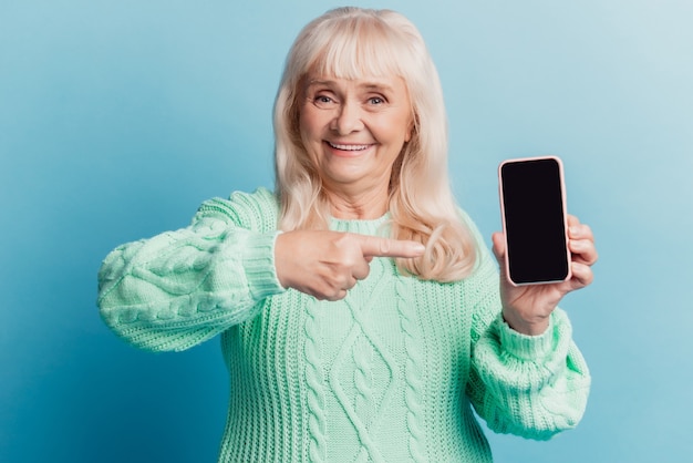 Positive old woman hold smartphone point finger touch screen isolated on blue background