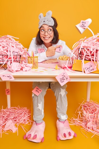 Positive optimistic brunette Asian woman in nightwear works at office desk surrounded by cut paper points away eats delicious cornflakes isolated over yellow wall