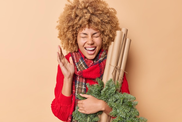 Positive overjoyed curly woman keeps hand raised exclaims loudly enjoys New Year preparations holds rolled paper and green wreath made of spruce branches wears scarf aound neck poses indoor.