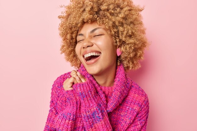 Positive overjoyed woman laughs out loudly keeps hands together\
has eyes closed dressed in warm knitted jumper chuckles over\
something funny poses against pink background genuine\
happiness