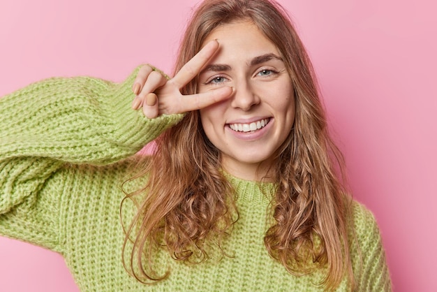 Positive peaceful blue eyed pretty woman with long wavy hair makes peace gesture over eye smiles gladfully has good mood wears green sweater isolated over pink background. Body language concept