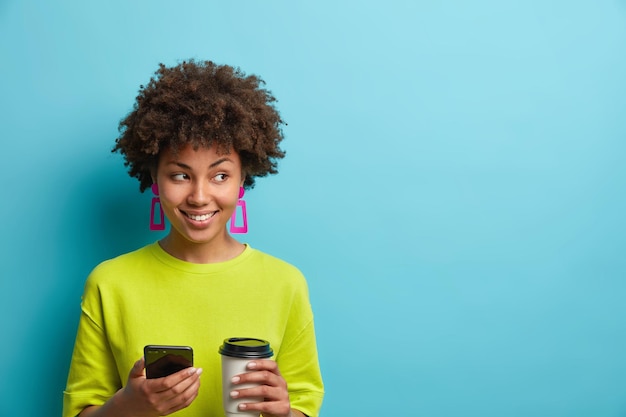 Positive pleasant looking woman holds smartphone and coffee to\
go scrolls newsfeed and enjoys favorite drink wears casual t shirt\
focused away isolated over blue background empty space for your\
text