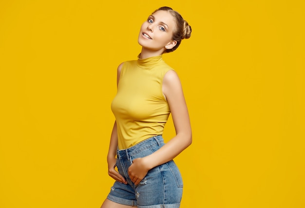 Positive portrait of gorgeous blonde hipster woman model with curly hair posing on colorful yellow background in studio