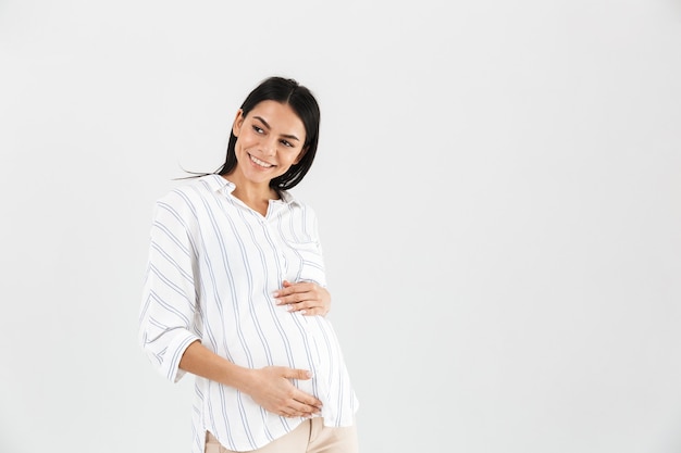 positive pregnant woman smiling and touching her big tummy while standing isolated over white wall