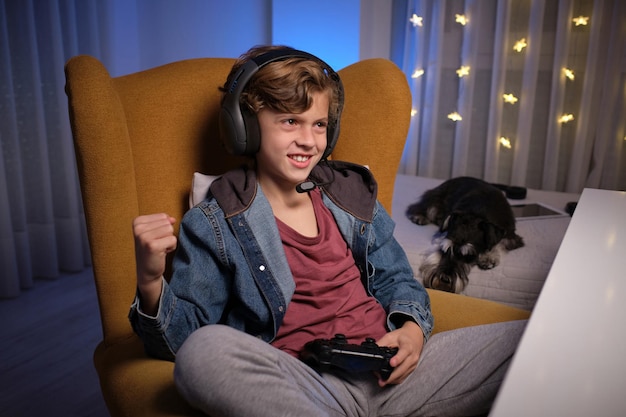 positive preteen boy in headset sitting on comfortable armchair with gamepad and celebrating achievement in videogame near bed with dog